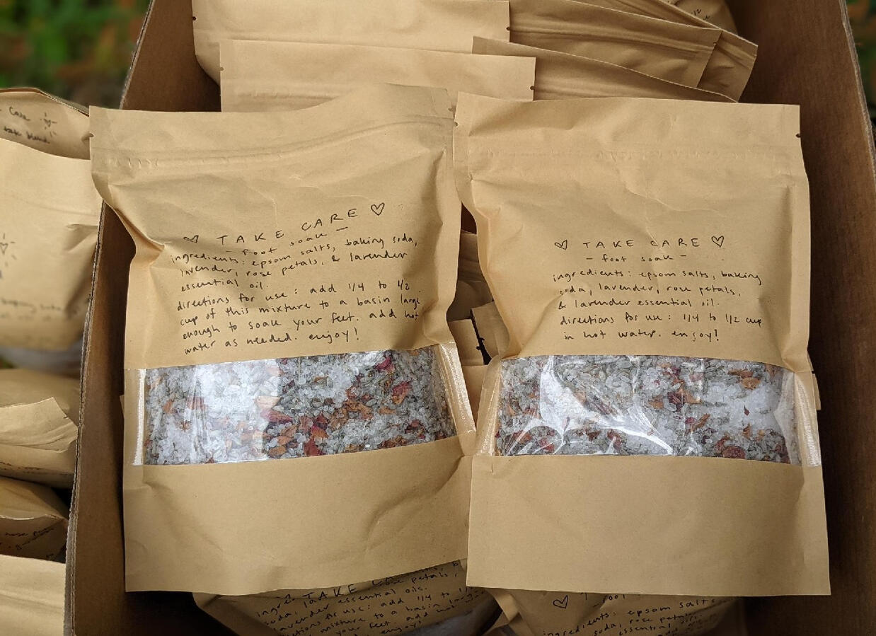 A box of paper bags of herbal bath salts that have a little plastic viewing window to see the contents, labeled "take care foot soaks"