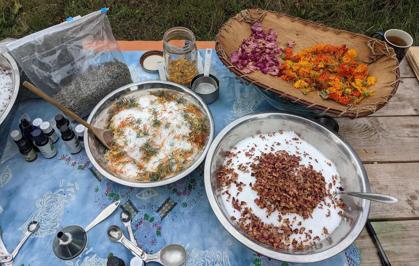 A work table outside with two bowls of epsom salts and herbs, a basket with dried calendula and rose petals, several bottles of essential oils, a bag of dried herbs and different measuring utensils