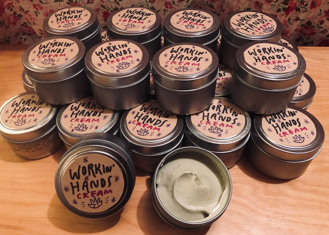 A stack of large tins labeled "workin' hands cream". One tin is open to show a smooth looking cream of light beige color